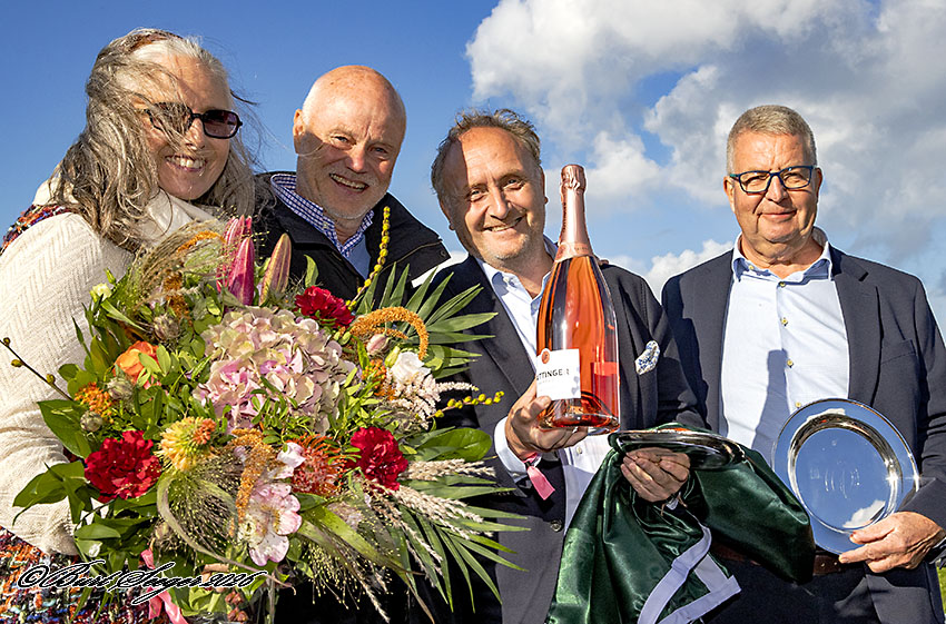 Fire personer med blomsterbuket, champagneflaske og sølvfad på sejrspodiet efter Dansk Oaks på Klampenborg i solskin.